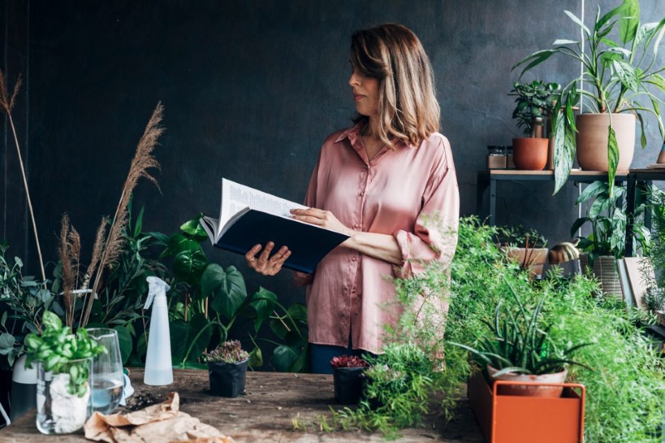 A woman paging through a gardening book, surrounded by various plants and potted plants.